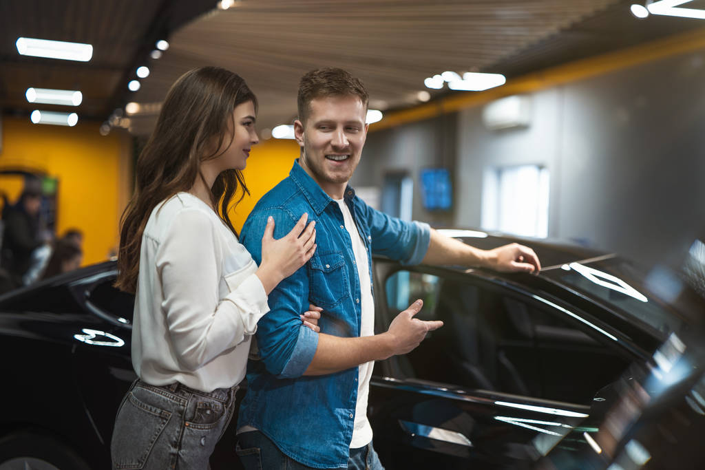 young couple handsome man and beautiful woman looking for new vehicle in electric car dealership center - Photo, Image