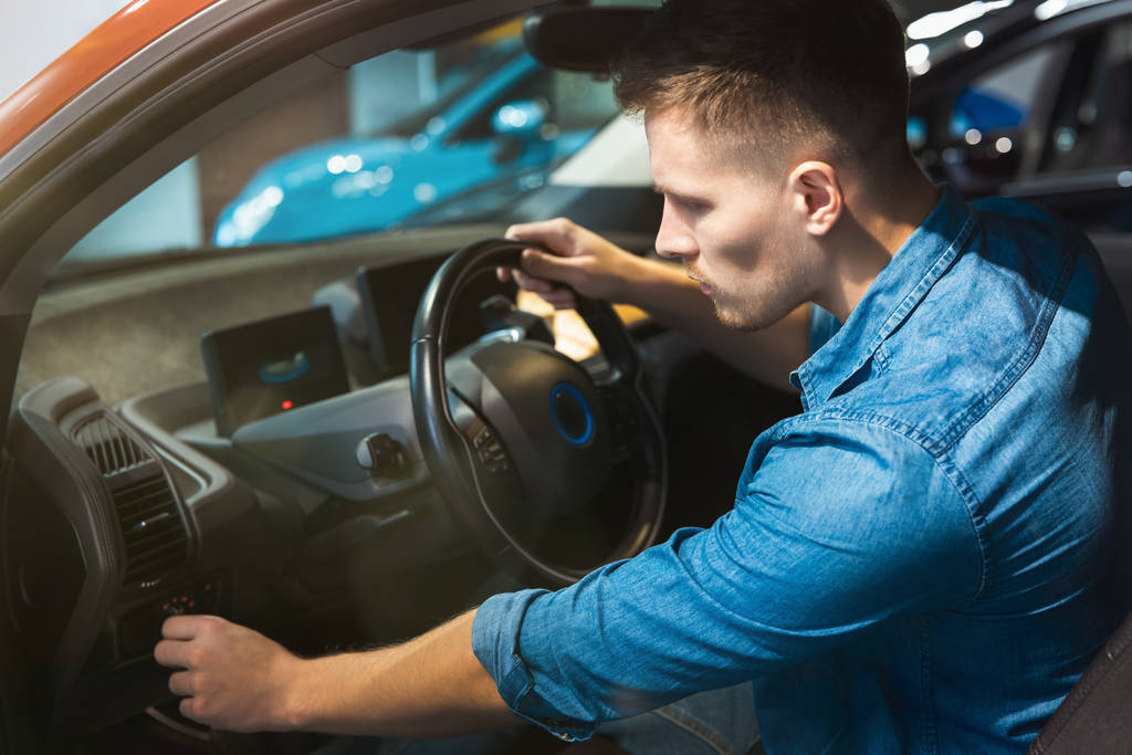 mladý pozitivní muž zákazník sedí v novém vozidle objevování interiéru v autosalonu, koncept nulových emisí - Fotografie, Obrázek