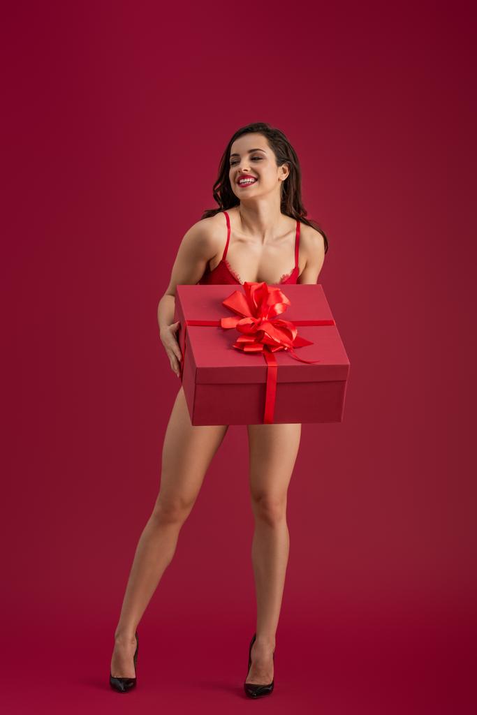 sexy meisje in lingerie en hoge hakken schoenen houden grote gift box terwijl weg te kijken en glimlachen op rode achtergrond - Foto, afbeelding