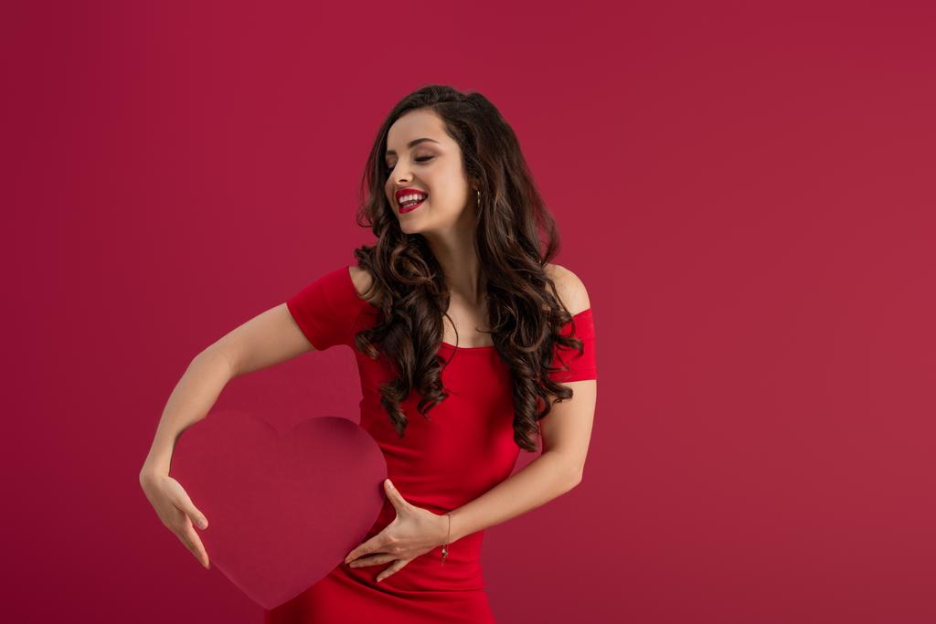 sexy, elegant meisje met papieren hart terwijl glimlachen met gesloten ogen geïsoleerd op rood - Foto, afbeelding