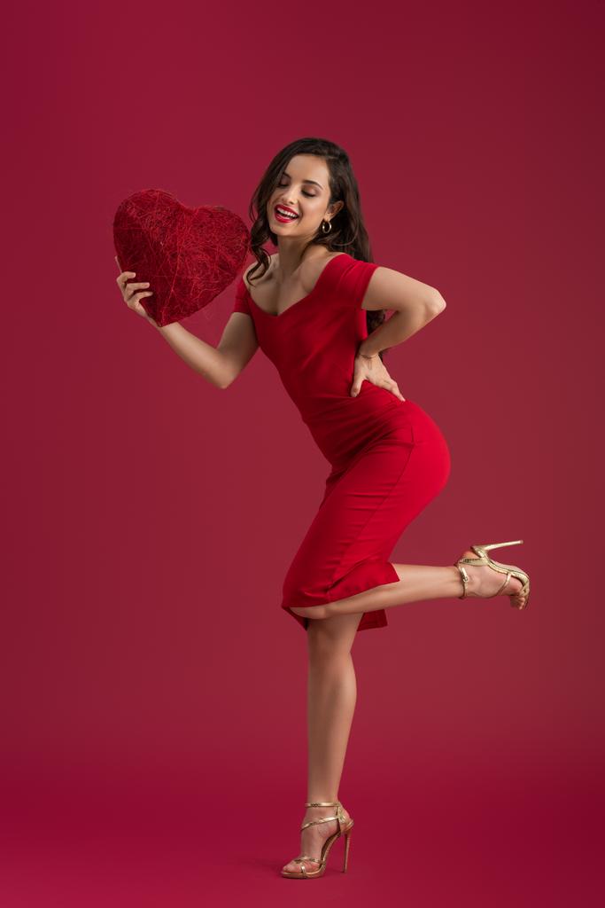 usmívající se, elegantní dívka drží dekorativní srdce, zatímco stojí na jedné noze na červeném pozadí - Fotografie, Obrázek