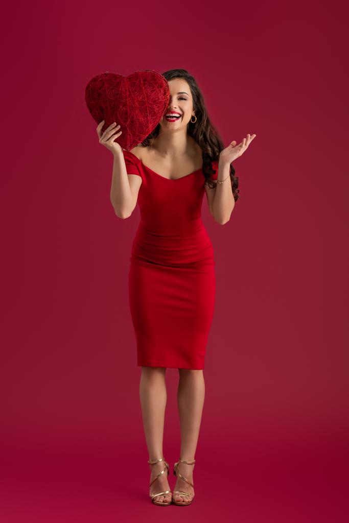 sorridente, menina elegante cobrindo os olhos com coração decorativo, enquanto em pé sobre fundo vermelho
 - Foto, Imagem