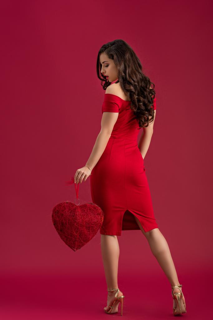 uwodzicielska, elegancka dziewczyna trzymająca dekoracyjne serce stojąc na czerwonym tle - Zdjęcie, obraz