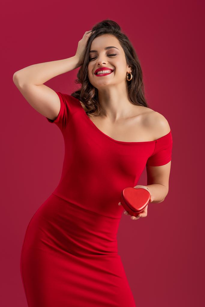 šťastná, elegantní dívka dotýkající se vlasů a drží srdce ve tvaru dárkové krabice, zatímco stojí se zavřenýma očima izolované na červené - Fotografie, Obrázek