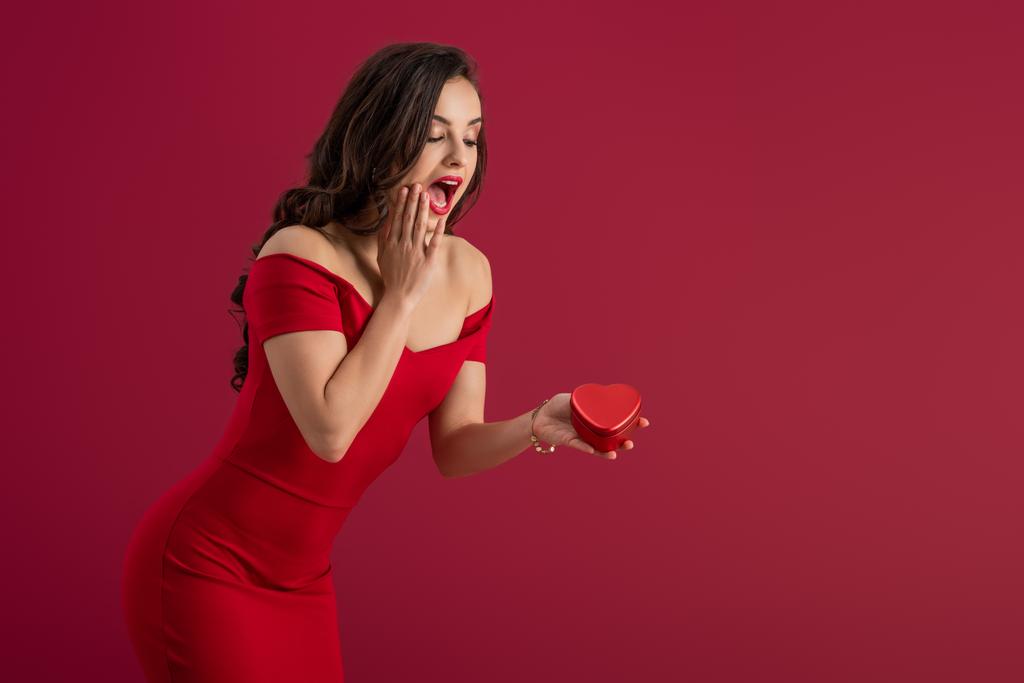 удивленный, элегантный девушка трогательное лицо, держа сердце форме подарочной коробки изолированы на красный
 - Фото, изображение