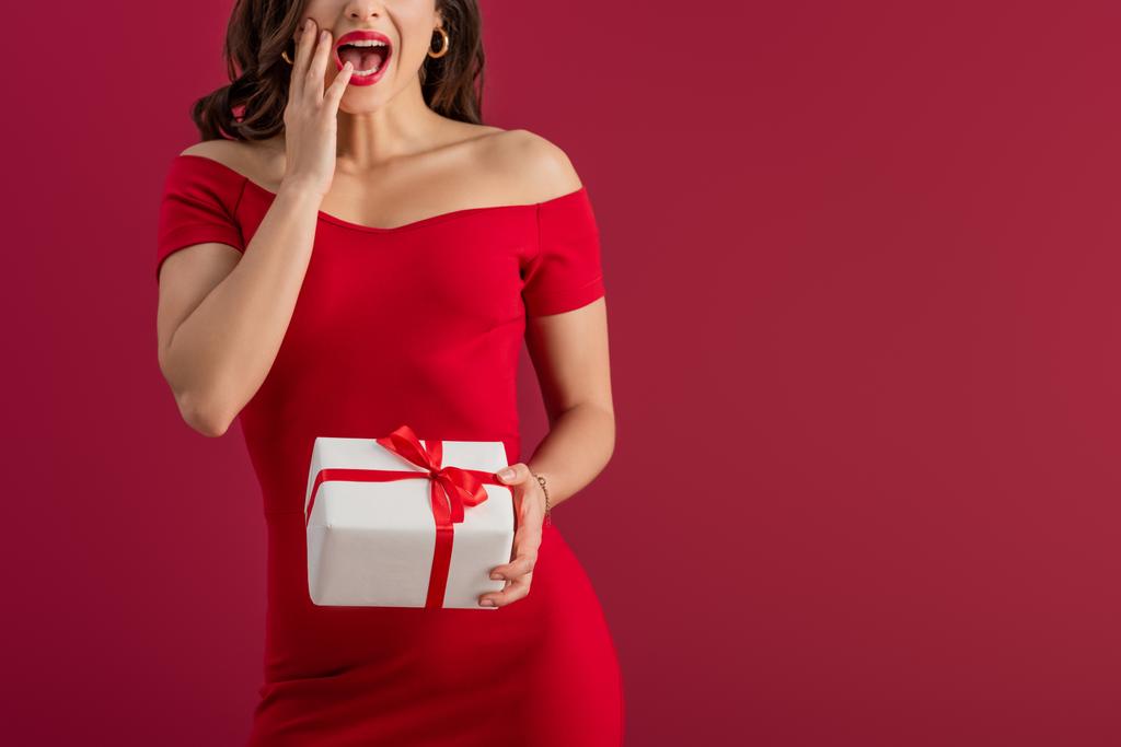 περικοπή άποψη του σοκαρισμένος, κομψό κορίτσι κρατώντας κουτί δώρου, ενώ αγγίζοντας το πρόσωπο απομονώνονται σε κόκκινο - Φωτογραφία, εικόνα