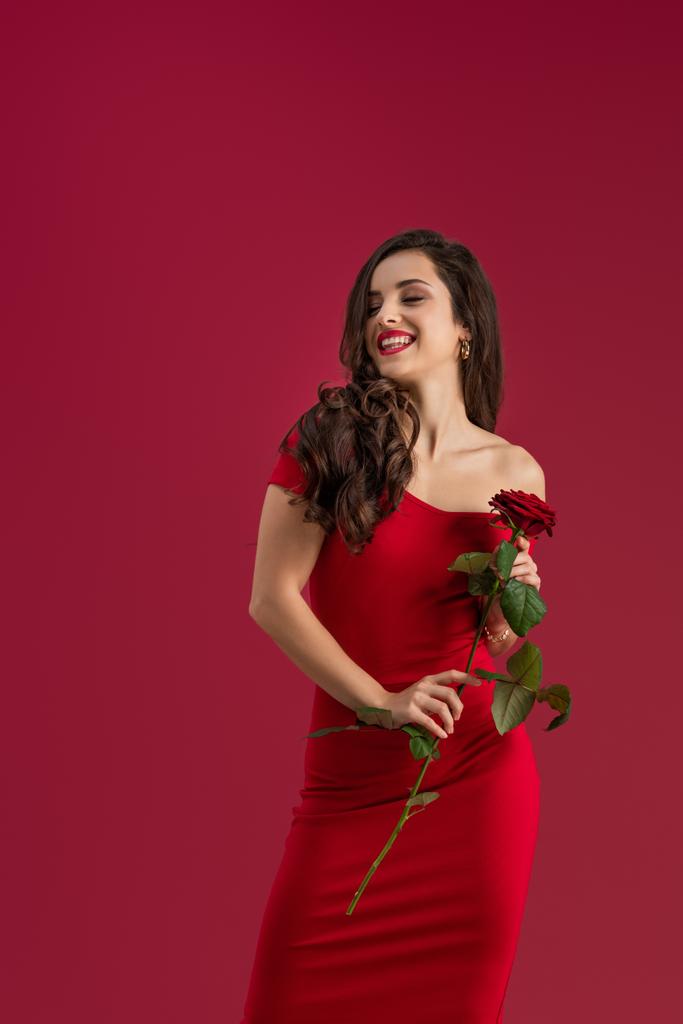 sexy, šťastná dívka drží červenou růži a usmívá se zavřenýma očima izolované na červené - Fotografie, Obrázek