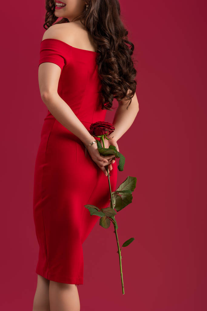terug uitzicht van sexy, elegant meisje met rode roos en glimlachen geïsoleerd op rood - Foto, afbeelding