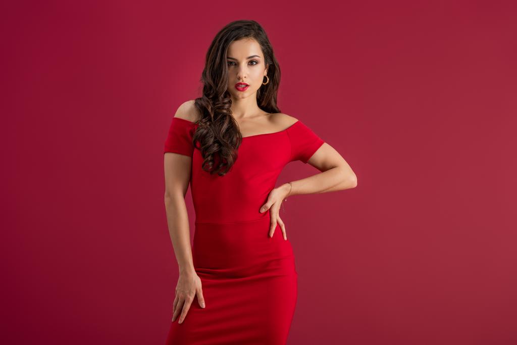 menina sedutora em vestido elegante olhando para a câmera enquanto posando com a mão no quadril isolado no vermelho
 - Foto, Imagem