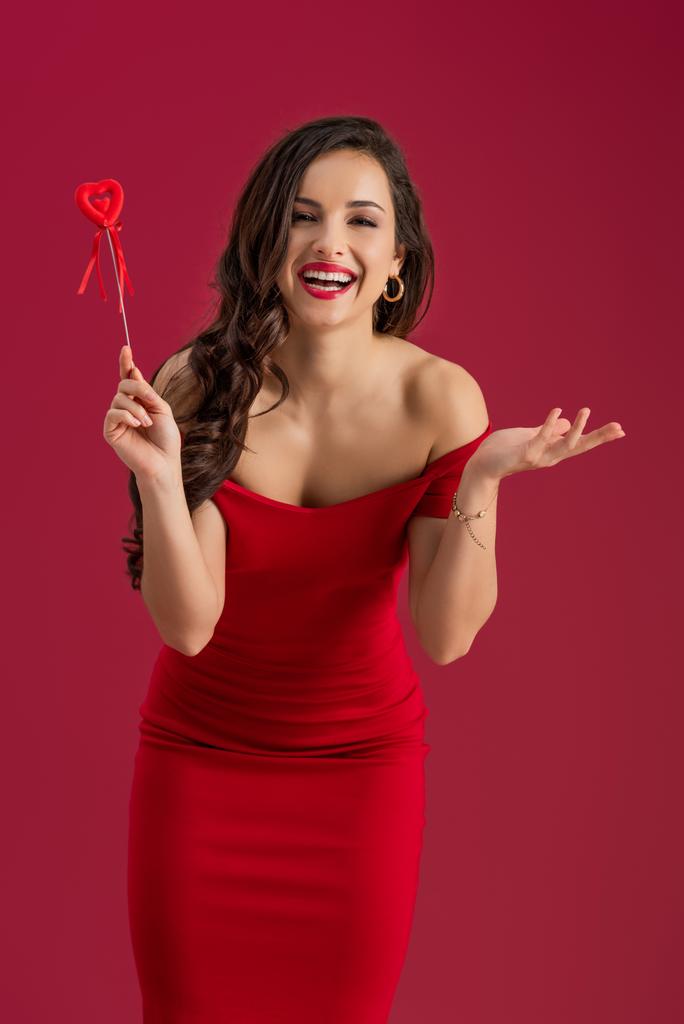 šťastná, elegantní dívka se usmívá na kameru, zatímco drží hračky srdce na hůl izolované na červené - Fotografie, Obrázek