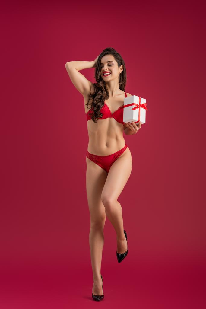 seductora chica en lencería y zapatos de tacón alto sosteniendo caja de regalo blanca y tocando el cabello con los ojos cerrados sobre fondo rojo
 - Foto, imagen