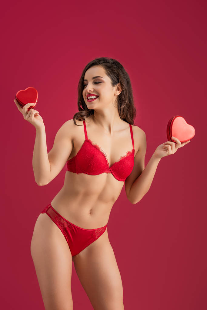 sexy, smiling girl in lingerie holding heart-shaped gift boxes isolated on red - Photo, Image