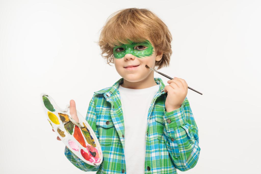 niño alegre con máscara de geco pintado en la cara tocando mejilla con pincel aislado en blanco
 - Foto, Imagen