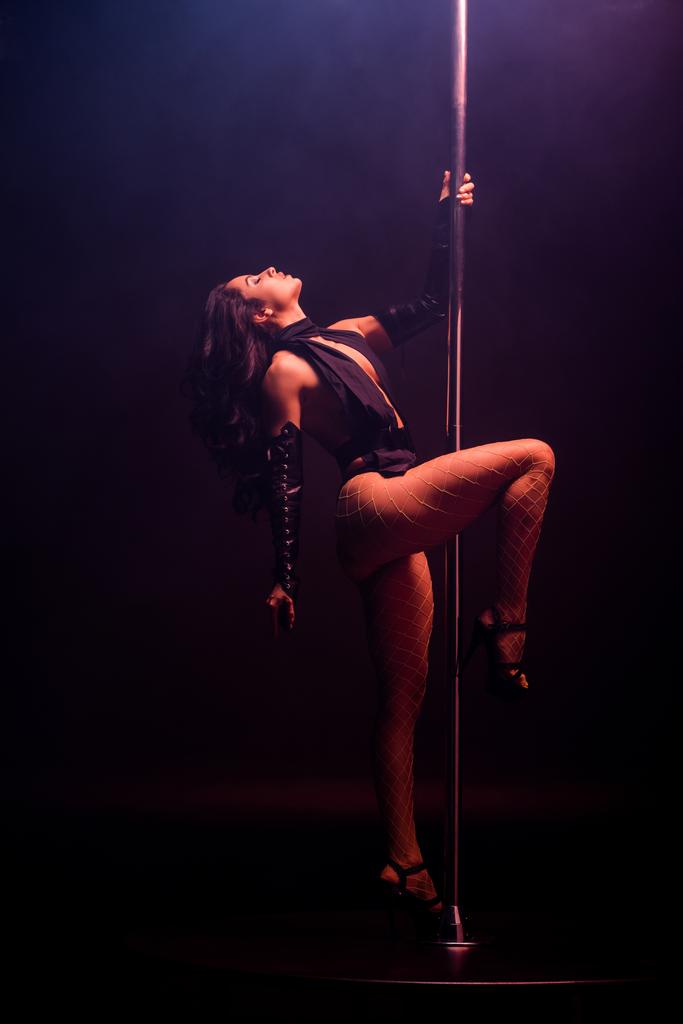 vista lateral de stripper quente dançando perto de pilão em preto com fumaça
  - Foto, Imagem