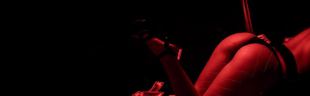 panoramic shot of passionate woman dancing striptease near dollar banknotes isolated on black with red lighting  - Photo, Image