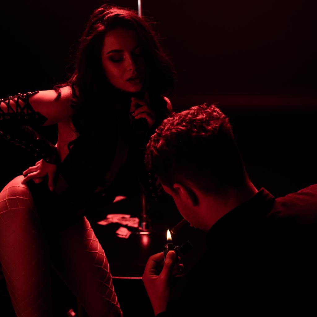 hombre sosteniendo más ligero mientras fuma cerca de stripper sexy en negro con iluminación roja
  - Foto, Imagen