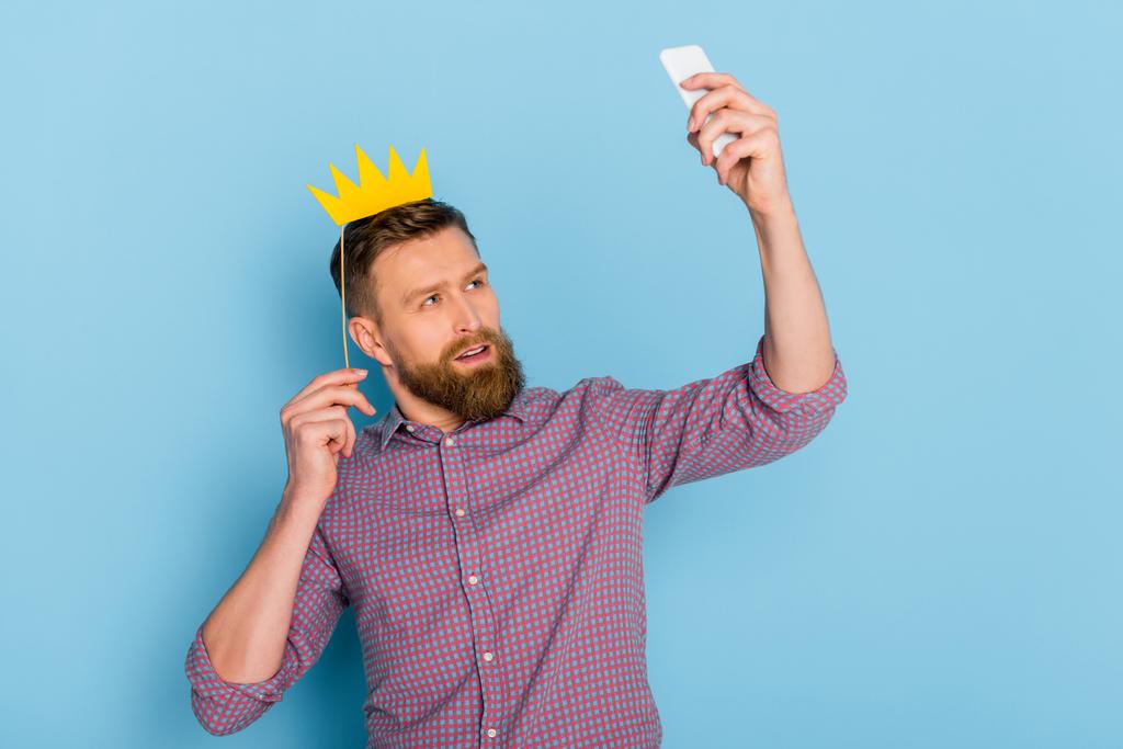 man met papieren kroon en het nemen van selfie op blauwe achtergrond  - Foto, afbeelding