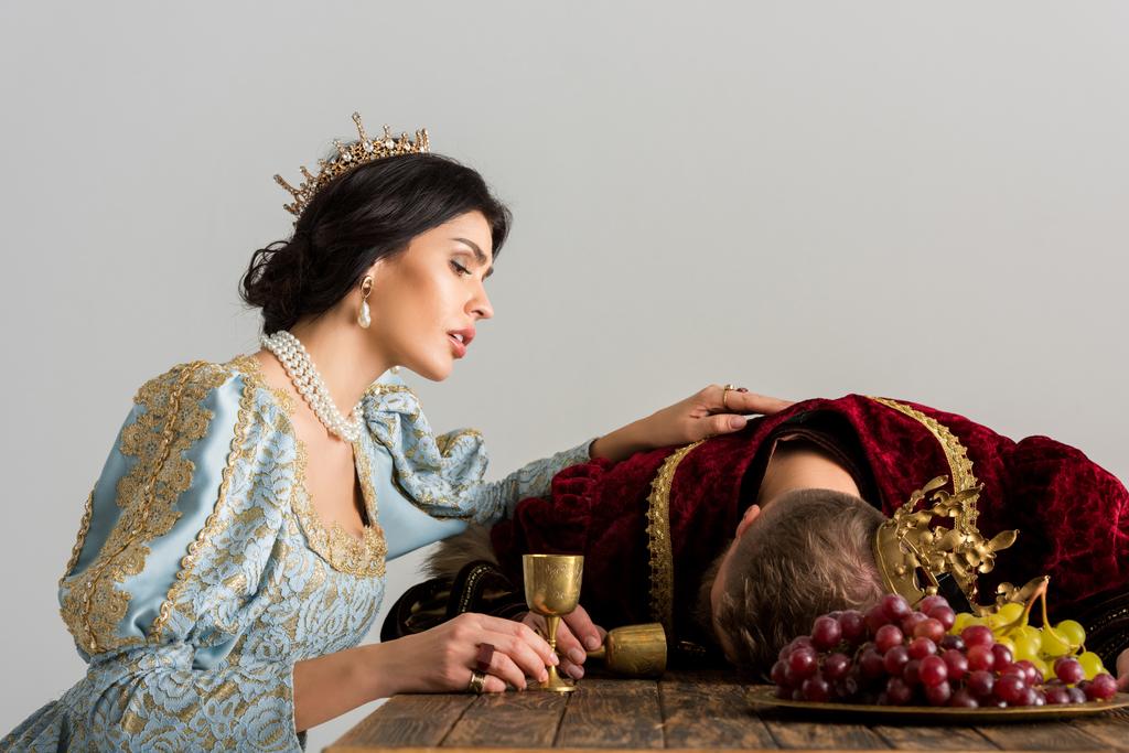 θλιμμένη βασίλισσα με το στέμμα να κοιτάζει τον νεκρό βασιλιά απομονωμένο στο γκρι - Φωτογραφία, εικόνα