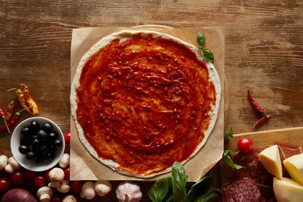 Вид пиццы на пергаментную бумагу с базиликом, овощами, оливками, ветчиной, салями, грибами и сыром на деревянном фоне
 - Фото, изображение