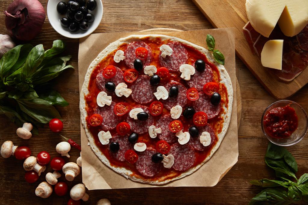 Vista superior da pizza com salame, cogumelos, azeitonas, molho de tomate, legumes, parmesão e presunto em fundo de madeira
 - Foto, Imagem