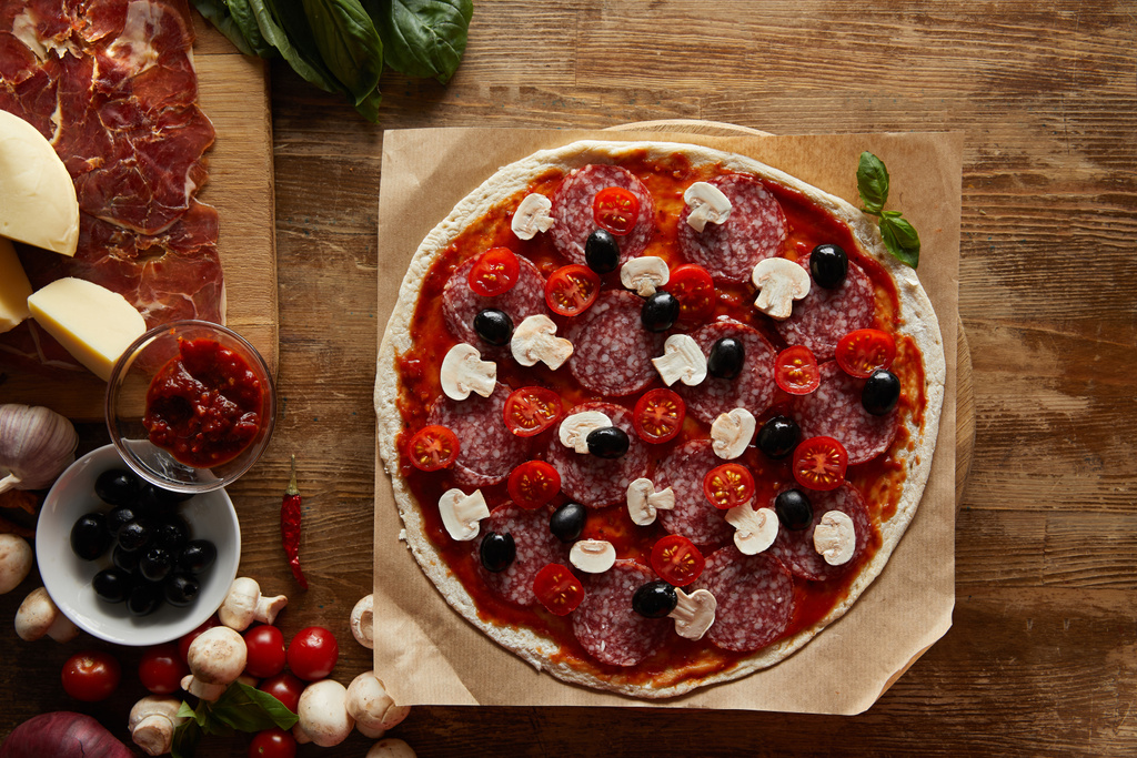 Bovenaanzicht van pizza met salami, champignons, olijven, tomatensaus, parmezaanse kaas, kerstomaten en prosciutto op houten ondergrond - Foto, afbeelding