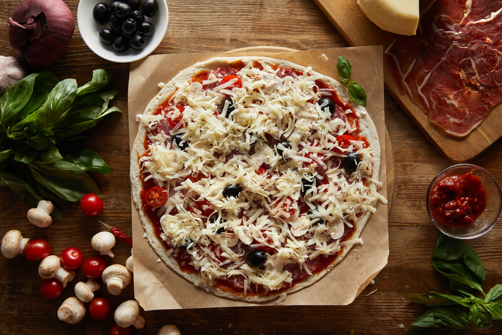 Vista superior da pizza em pergaminho com parmesão, legumes, azeitonas e salame em fundo de madeira
 - Foto, Imagem