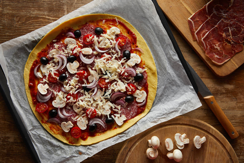 Top view of delicious pizza on parchment paper, mushrooms and prosciutto on wooden background - 写真・画像