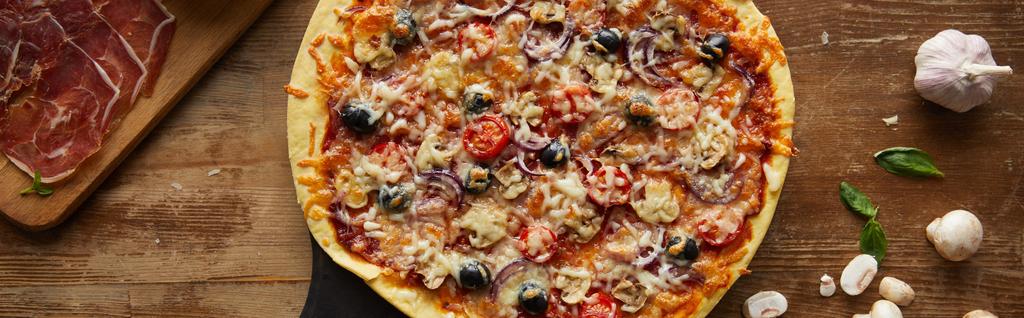 Horní pohled na chutnou pizzu a česnek, prosciutto, houby a bazalka na dřevěném pozadí, panoramatický výstřel - Fotografie, Obrázek