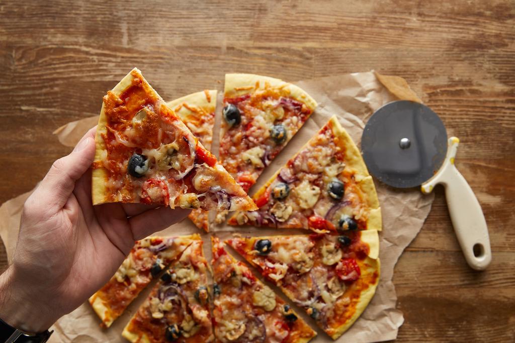 abgeschnittene Ansicht eines Mannes, der ein Stück Pizza auf hölzernen Hintergrund hält - Foto, Bild
