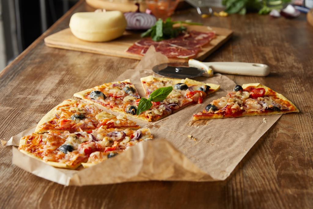selectieve focus van heerlijke Italiaanse pizza met olijven op bakpapier in de buurt van ingrediënten en pizza mes op houten tafel - Foto, afbeelding