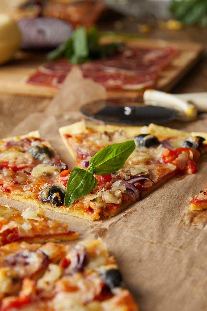 избирательный фокус вырезанной итальянской пиццы с оливками на выпечке бумаги рядом с ингредиентами и нож для пиццы на деревянном столе
 - Фото, изображение