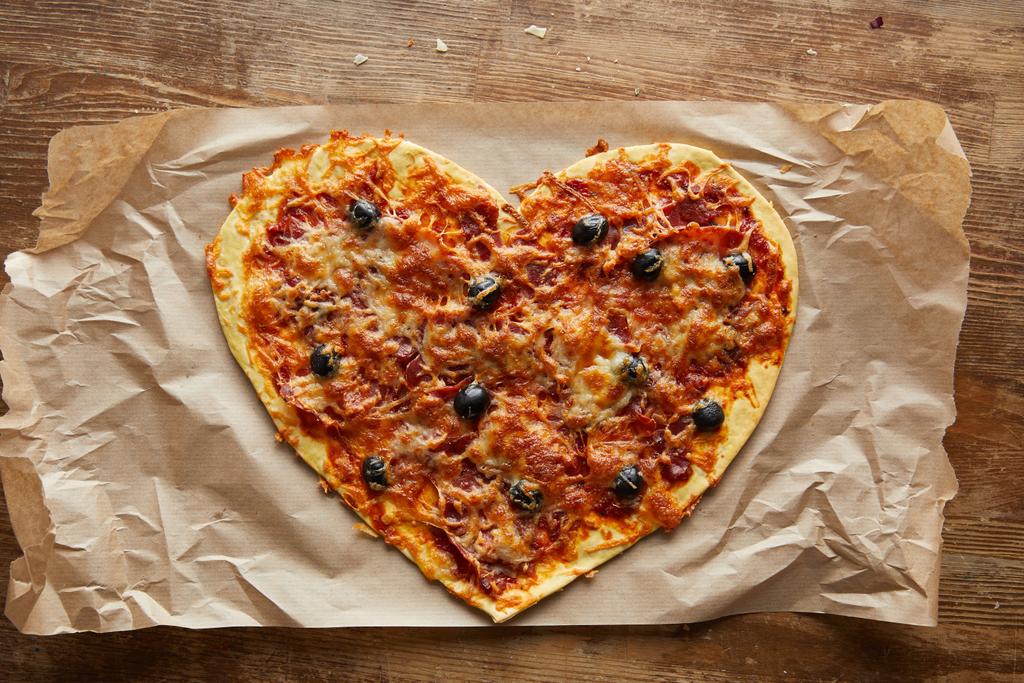 vrchní pohled na lahodnou italskou pizzu ve tvaru srdce na pečicím papíru na dřevěném stole - Fotografie, Obrázek