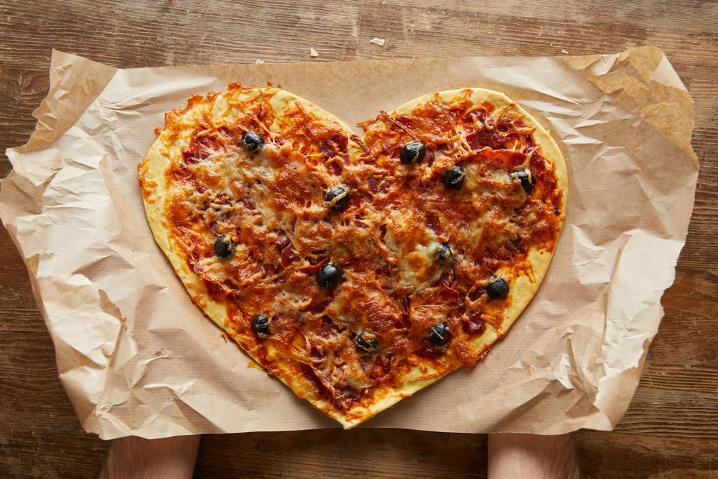 Kalbinde lezzetli İtalyan pizzası taşıyan bir adam ahşap masada kağıt pişiriyor. - Fotoğraf, Görsel
