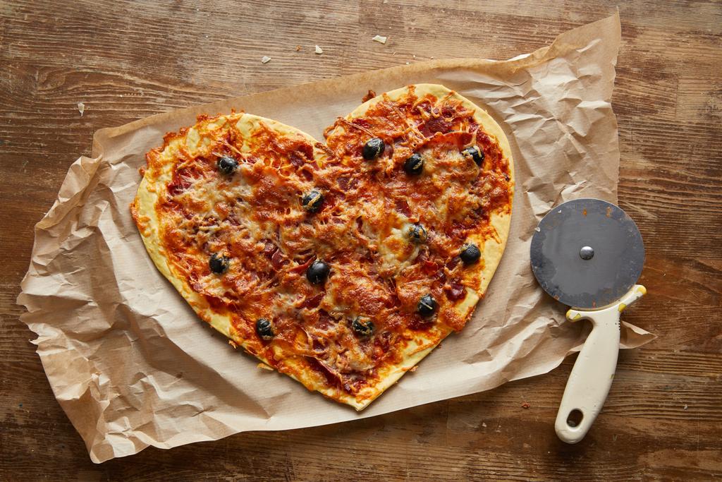 вид зверху смачної італійської піци у формі серця на папері для випічки біля піцерійного ножа на дерев'яному столі
 - Фото, зображення