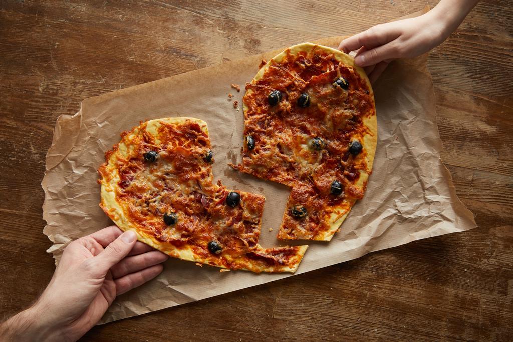 木製のテーブルの上で紙を焼く上で壊れた心の形でおいしいイタリアのピザを保持カップルのクロップドビュー - 写真・画像