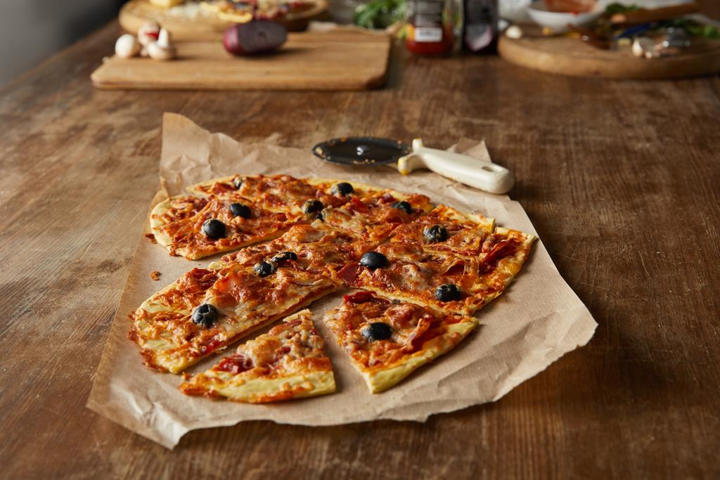 leckere italienische Pizza in Herzform in Stücke geschnitten auf Backpapier in der Nähe von Pizzamesser und Zutaten auf Holztisch - Foto, Bild