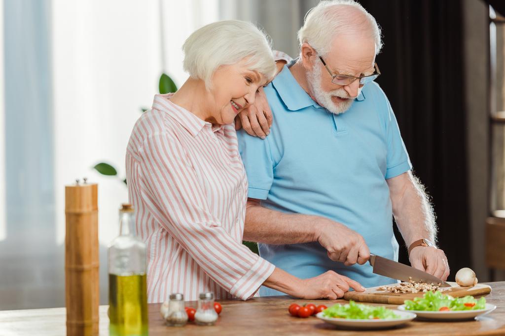 Χαμογελαστή ηλικιωμένη γυναίκα στέκεται δίπλα στον άντρα της κόβοντας λαχανικά στο τραπέζι της κουζίνας  - Φωτογραφία, εικόνα