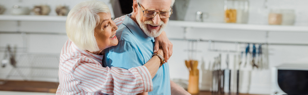 Панорамный снимок пожилой женщины, обнимающей улыбающегося мужа на кухне
 - Фото, изображение