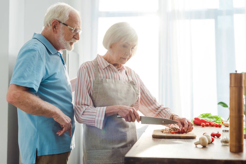 Вибірковий фокус старшого чоловіка, що стоїть за дружиною, що ріже свіжі овочі на кухонному столі
 - Фото, зображення
