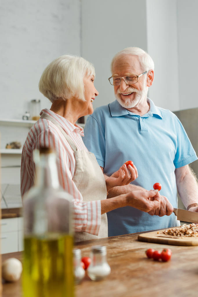 Mutfak masasında mantar keserken karısına kirazlı domateslerle bakan gülümseyen adamın seçici odağı - Fotoğraf, Görsel
