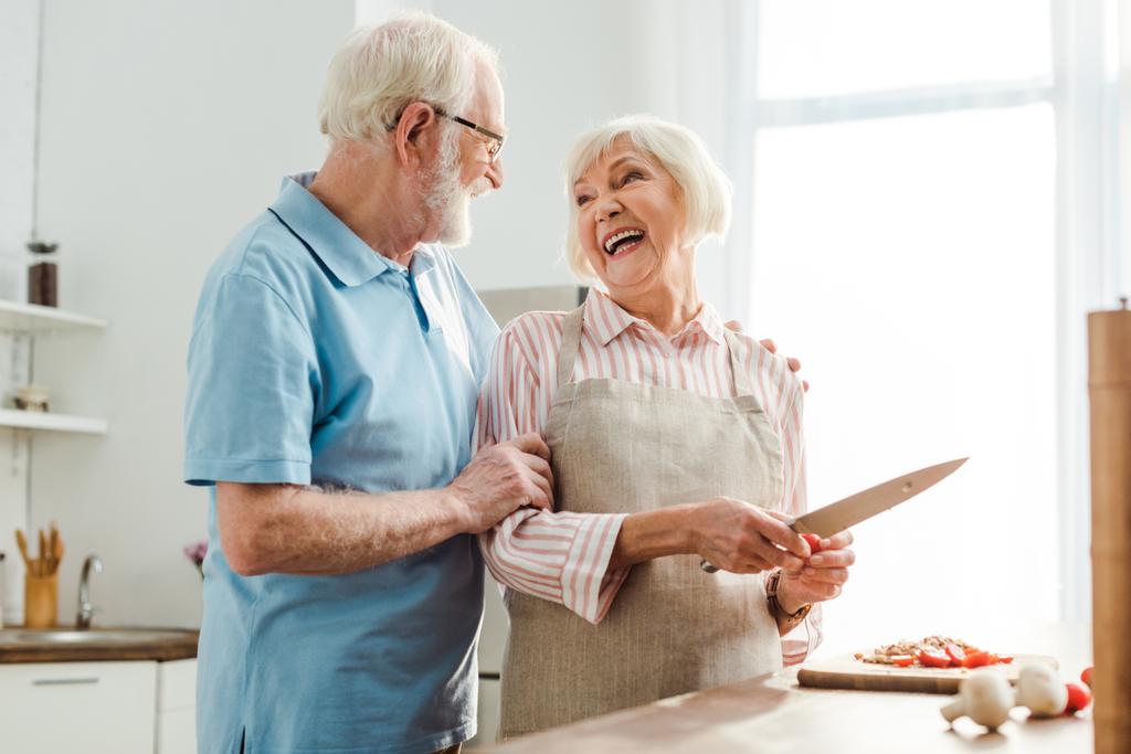 Ηλικιωμένος άντρας αγκαλιάζει χαμογελαστή γυναίκα ενώ κόβει λαχανικά στο τραπέζι της κουζίνας - Φωτογραφία, εικόνα