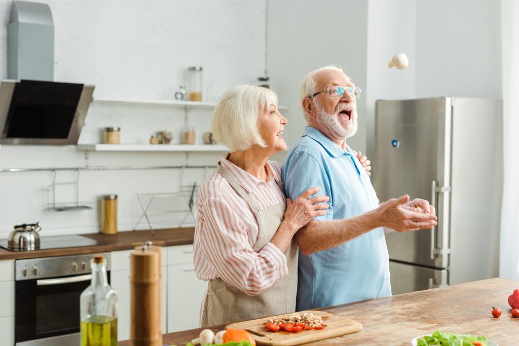 キッチンテーブルの上に新鮮な野菜でキノコを投げる男が笑っているシニアカップル - 写真・画像