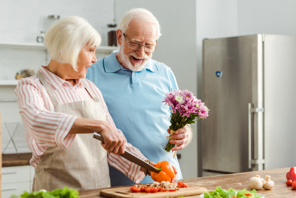 Senior man giving bouquet to surprised wife during cutting fresh vegetables in kitchen - Photo, Image