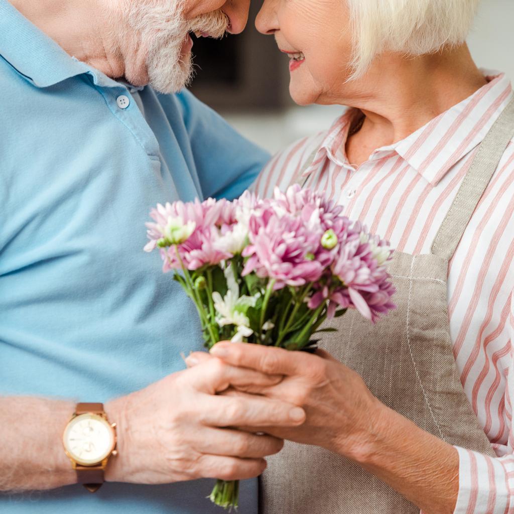 Vista cortada de casal de idosos sorrindo um para o outro e segurando buquê de crisântemos
 - Foto, Imagem