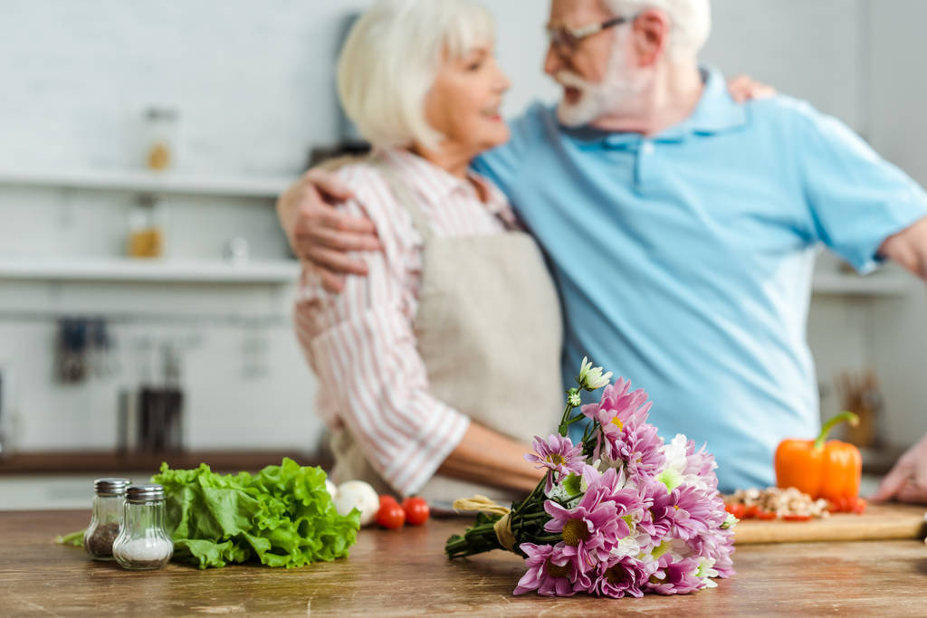 テーブルの上の花束と新鮮な野菜とキッチンの笑顔のシニアカップルの選択的な焦点 - 写真・画像