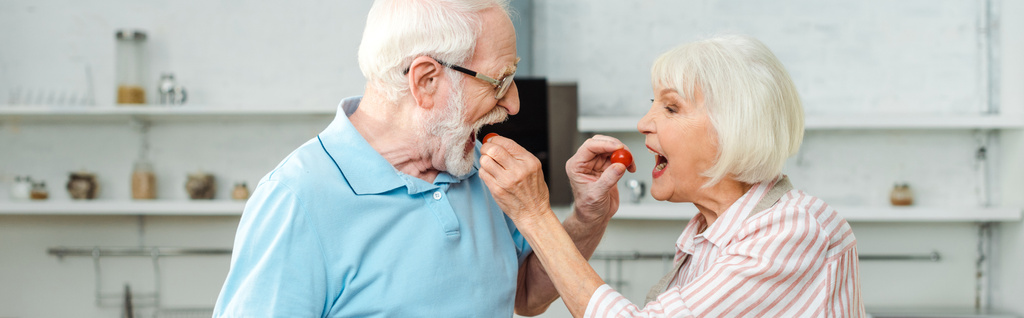Oldalnézet idős pár etetés egymást cseresznye paradicsom a konyhában, panoráma lövés - Fotó, kép