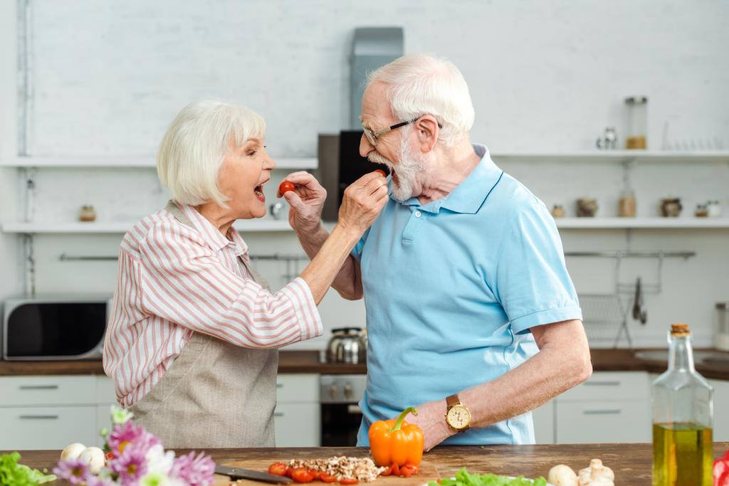 Vista lateral de pareja mayor alimentándose con tomates cherry mientras cocinan en la mesa de la cocina
 - Foto, imagen