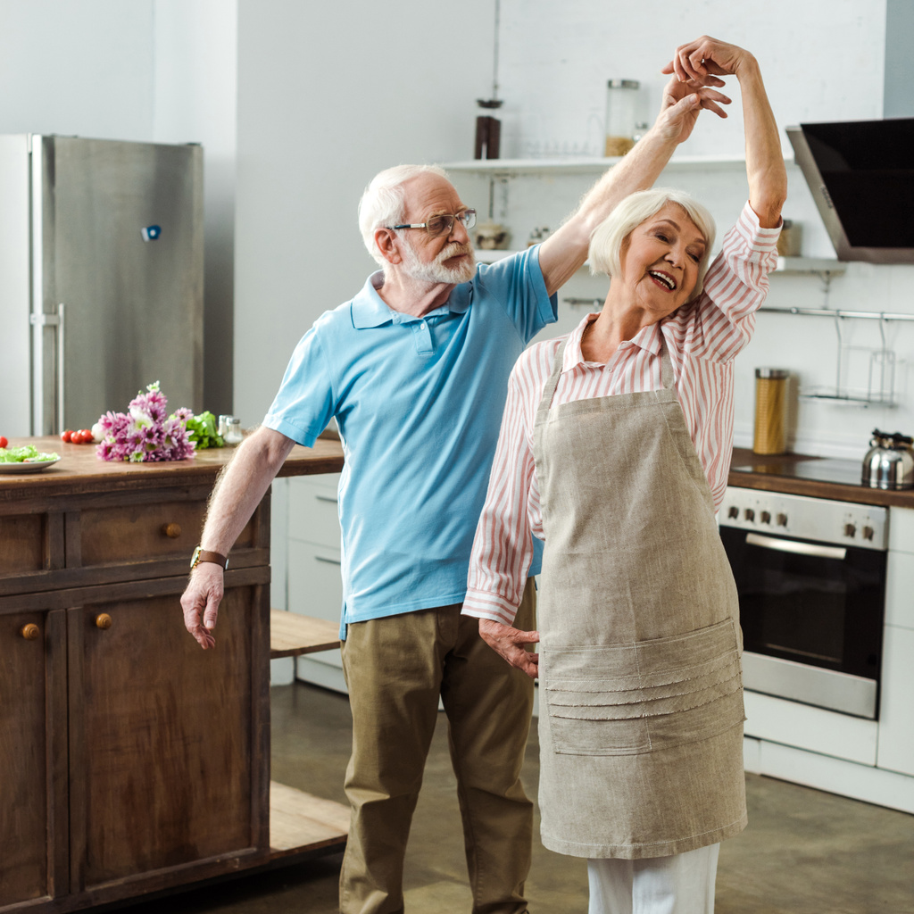 Seniorchef tanzt mit lächelnder Frau bei Gemüse und Strauß auf Küchentisch - Foto, Bild