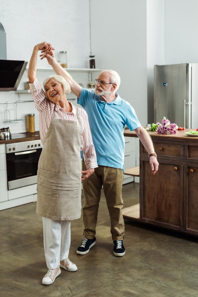 Idősebb férfi táncol mosolygós feleséggel kötényben a konyhában - Fotó, kép