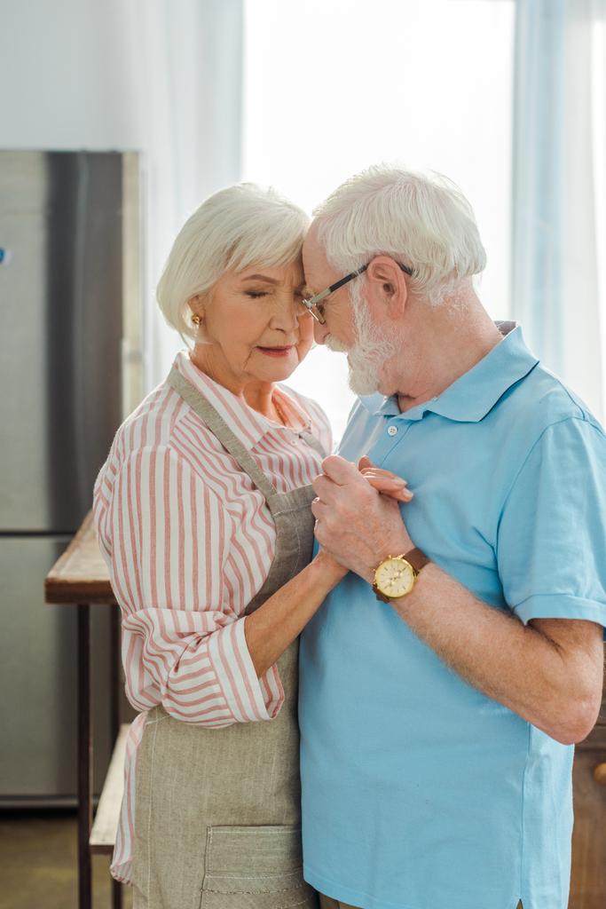 Vista lateral de una pareja de ancianos tomados de la mano mientras se abrazan en la cocina
  - Foto, Imagen